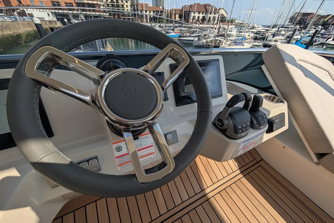 Boat helm with a steering wheel, control levers, and a display screen, overlooking a marina filled with yachts and boats.